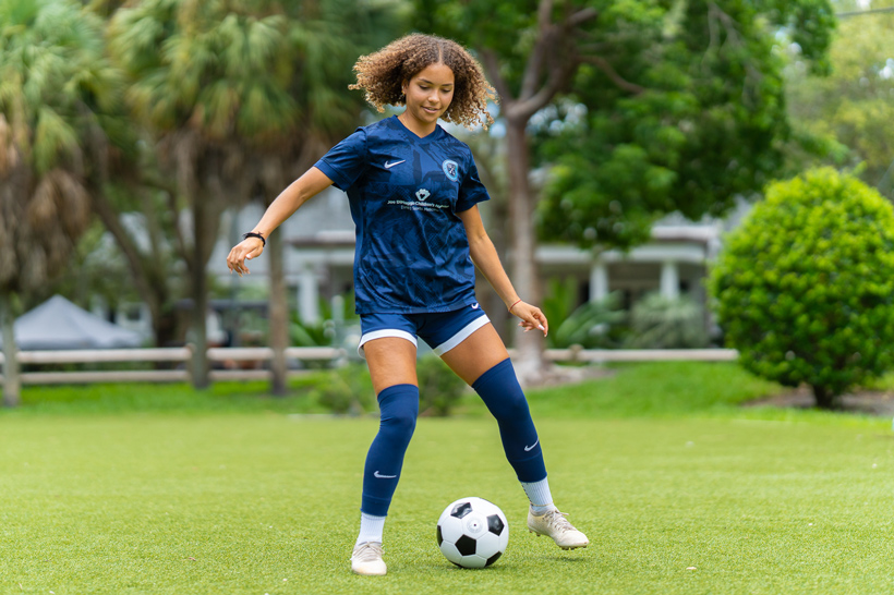 Young girl wearing blue jersey holding soccer ball in front of soccer field.